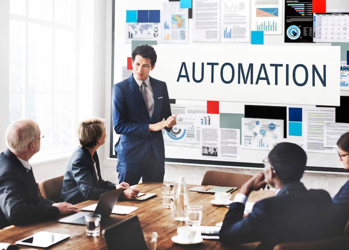A business meeting with a presenter standing by a board displaying charts and data related to automation, while colleagues listen attentively.