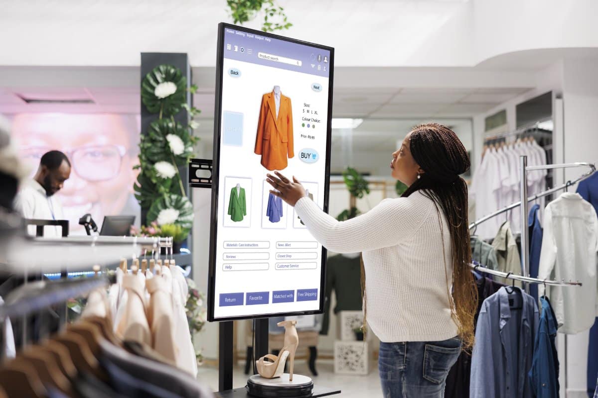 A shopper interacts with a touchscreen display showcasing clothing options, including an orange blazer, in a modern retail environment.